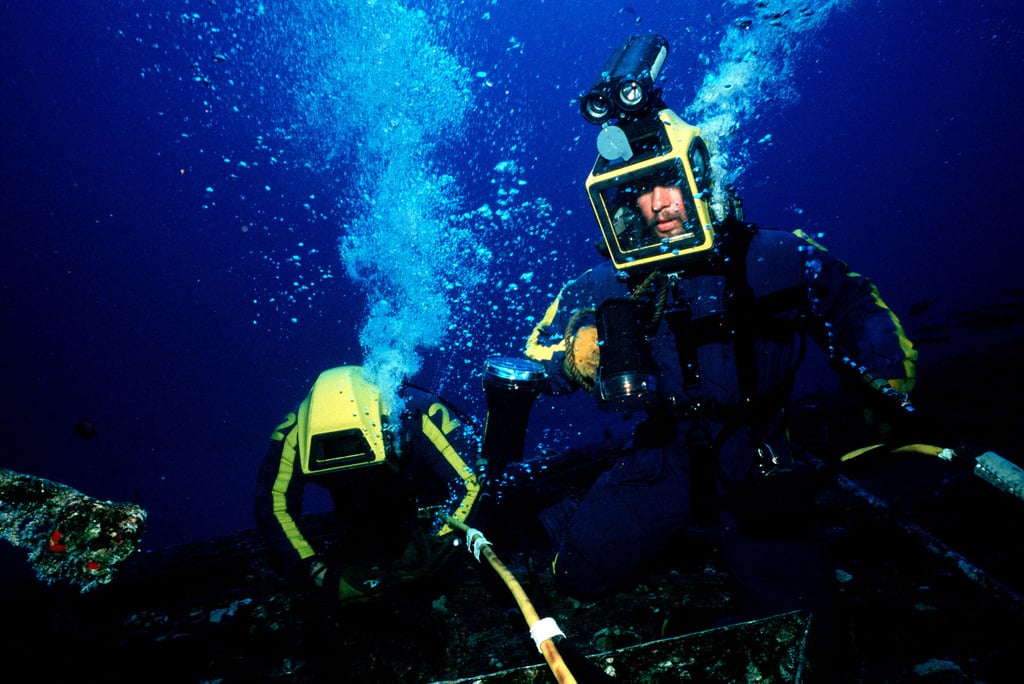 Two divers in yellow helmets working underwater with breathing equipment, surrounded by bubbles. A rope extends between them, set against a dark blue aquatic background.