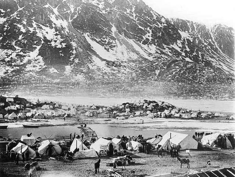 Black and white photo of a sprawling tent camp at the base of snow-capped mountains near a body of water. Numerous tents and small structures are visible, with people and animals moving around the foreground.