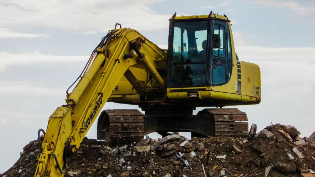 A yellow excavator sits atop a pile of rubble and dirt. Its arm is extended outward, and the sky in the background is partly cloudy.