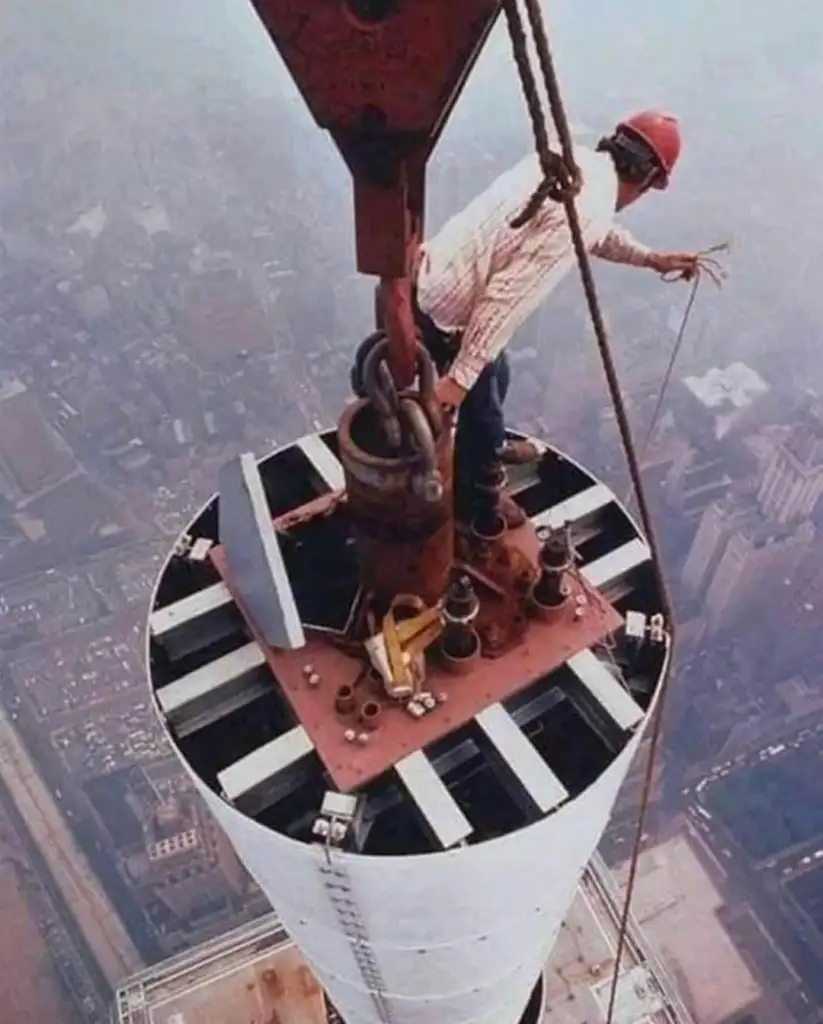 A construction worker wearing a red hard hat stands on top of a tall, cylindrical structure high above a cityscape. The worker is holding onto a cable attached to a large crane hook. The city below is hazy under a clear sky.