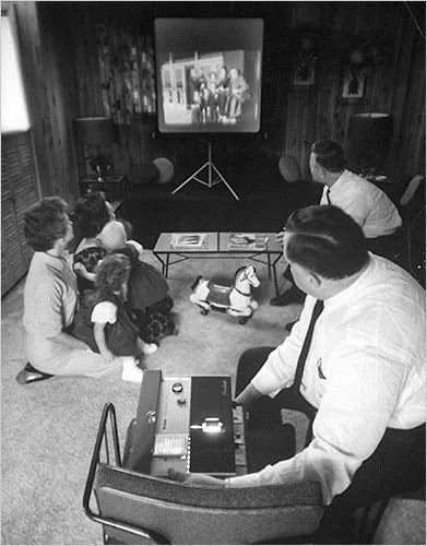 A vintage black and white photo shows a family gathered in a living room, watching a home movie projected onto a screen. Two adults and two children sit on the floor, while another adult operates the projector. A toy horse is on the carpet.