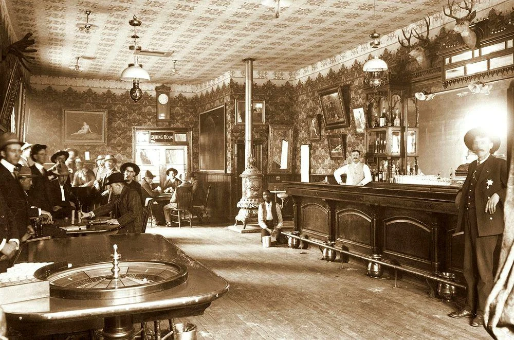 A vintage saloon scene with a bartender behind a long wooden bar, people in early 20th-century attire, and a roulette table in the foreground. The room has ornate wallpaper, mounted antlers, and a decorative stove.