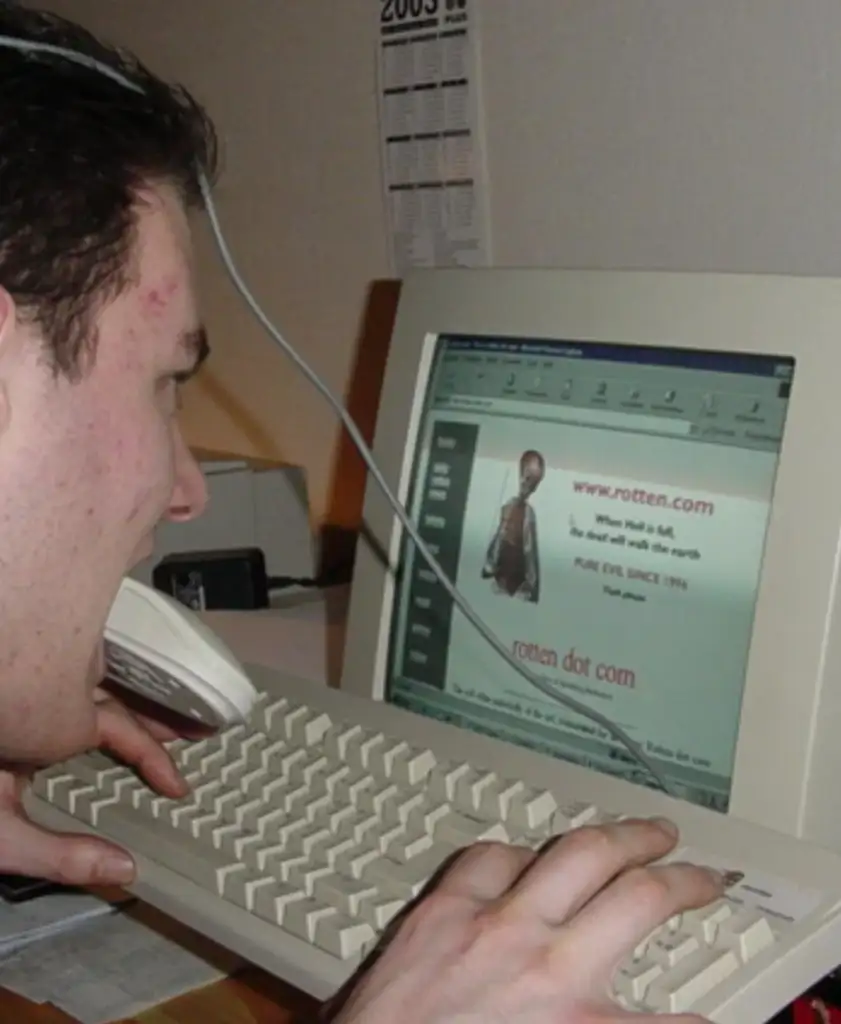 A person holds a telephone handset up to their mouth while using a computer featuring a website with "rotten dot com" on the screen. They are leaning close to the monitor, typing on a white keyboard.