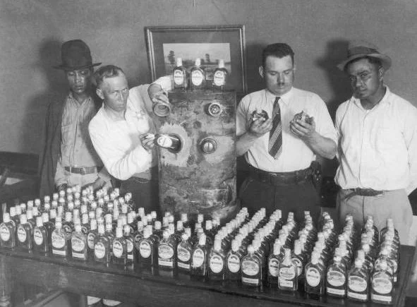 Four men stand behind a table filled with liquor bottles, examining labels and a large metal still. They appear to be participating in an inspection or investigation. The scene suggests an era of prohibition.