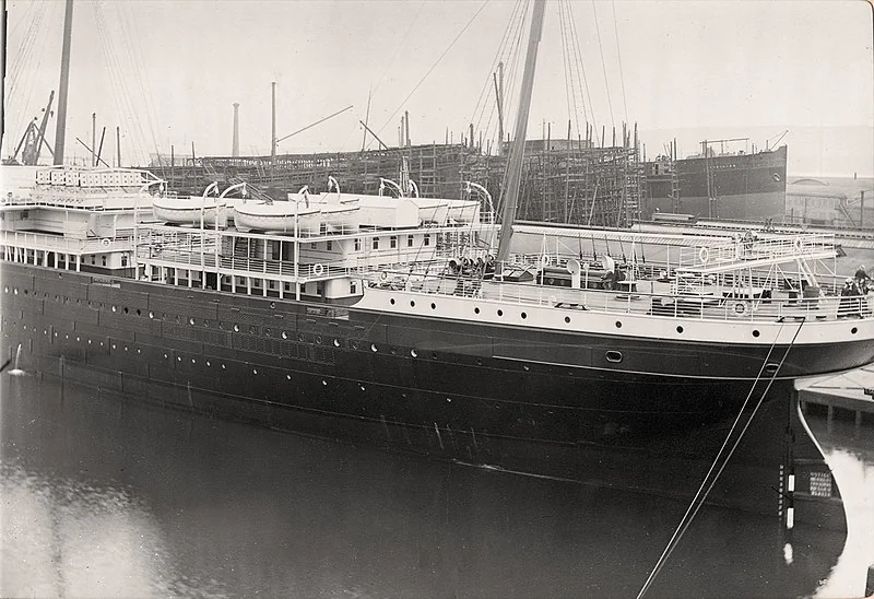 Black and white photo of a large ship docked at a harbor. The ship features multiple decks and lifeboats. In the background, there's a shipyard with cranes and another ship under construction.