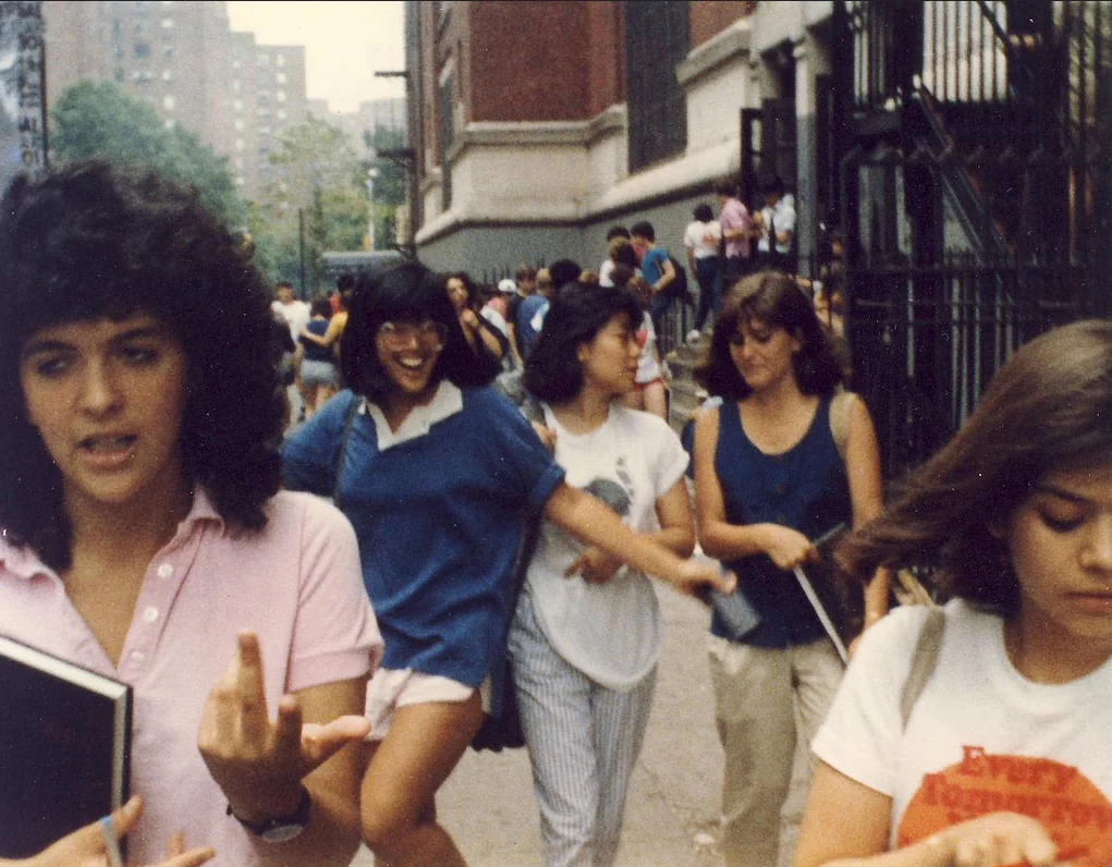 A group of young people walks outside a school building on a city street. One girl in the front has curly hair and holds a book. Others are smiling and laughing. The background shows trees, more students, and urban buildings.