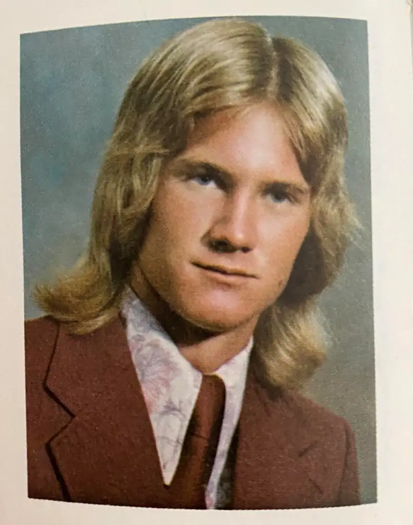 A young person with long, light brown hair wears a maroon suit jacket and a patterned collared shirt with a matching tie. The background is a soft blue-gray, typical of vintage portrait photos.