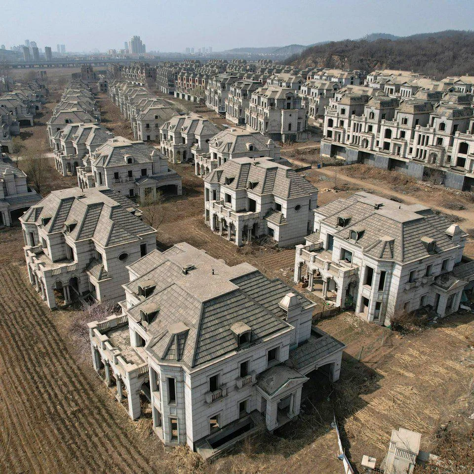 Aerial view of an abandoned housing development featuring numerous identical, large grey houses with gabled roofs and empty windows. The area is overgrown with dry grass and weeds, stretching into the distance under a clear sky.