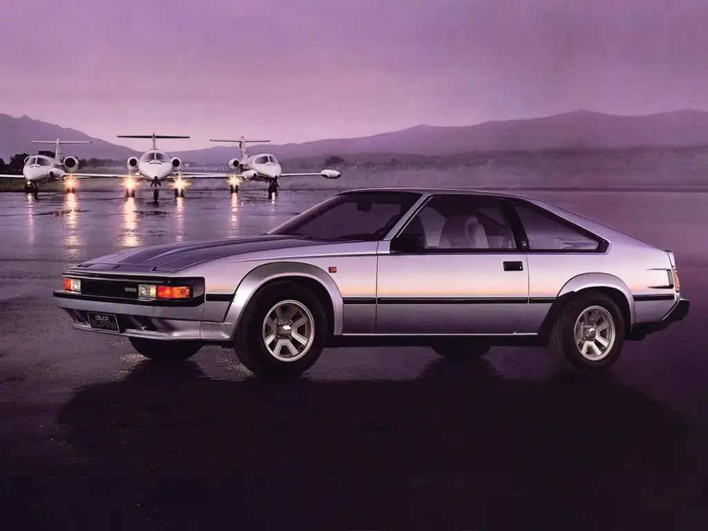 A silver sports car is parked on a wet tarmac with three small jets in the background. The sky is overcast, creating a dramatic and moody atmosphere. The scene is set against distant, silhouetted mountains.