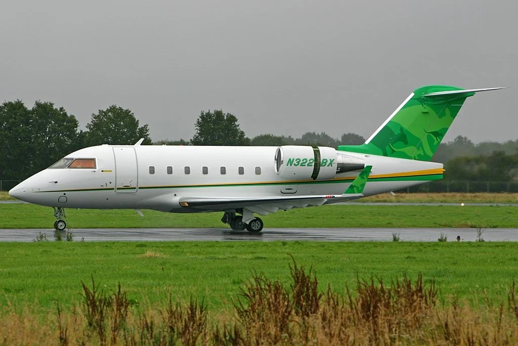 A small private jet with a white and green exterior, featuring the registration number N322GX, is taxiing on a wet runway. The background includes a grassy area and trees under an overcast sky.