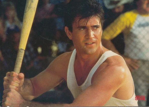 A man in a white tank top holds a baseball bat, poised to swing. He appears focused and is outdoors, with a blurred background of people and greenery.