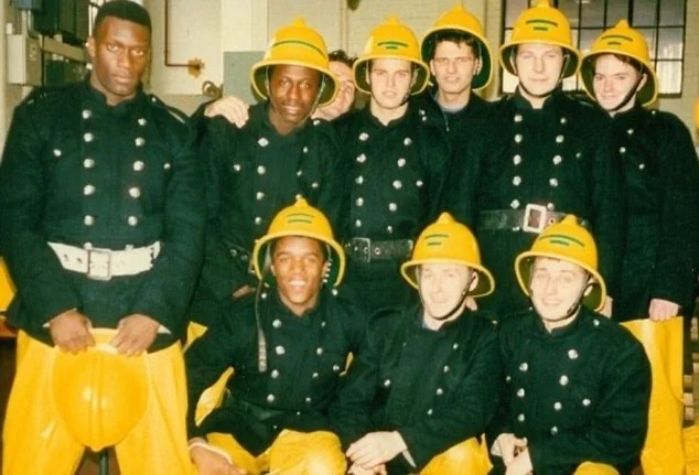 A group of firefighters pose indoors wearing vintage uniforms. They are dressed in black jackets with white buttons, yellow helmets, and yellow pants. Some are standing, while others are kneeling in front. The setting appears to be a fire station.