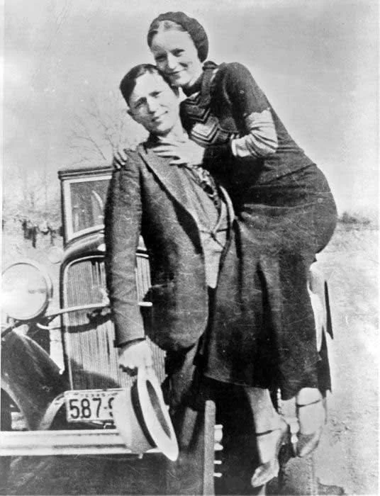 A woman in a dark dress sits on the front fender of a vintage car, leaning on a man in a suit who holds a hat in his hand. They both smile at the camera. The car has round headlights and a visible license plate number.