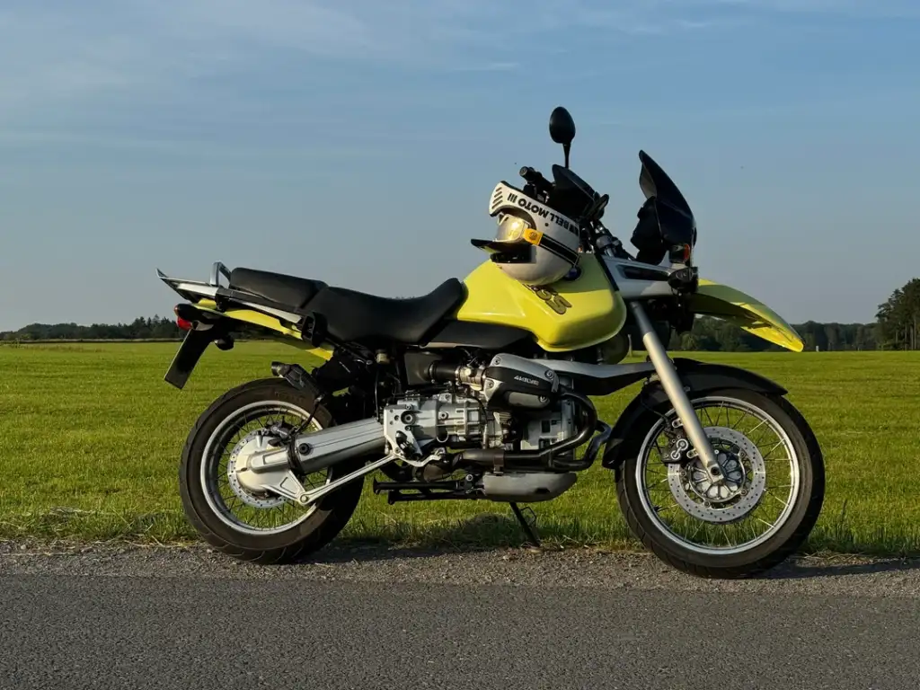 A yellow and black motorcycle is parked on the side of a rural road with green fields and trees in the background. A helmet is placed on the seat. The sky is clear and blue.