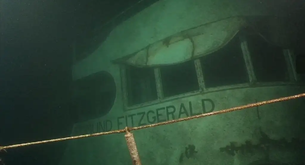 Underwater photo of the shipwreck of the Edmund Fitzgerald. The hull and windows are partially covered in algae. The ship's name is visible, and a rusted metal rail is in the foreground.