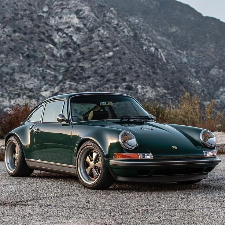 A vintage dark green sports car parked on a paved road in front of a mountainous backdrop. The car features classic round headlights, sleek curves, and retro styling, conveying a sense of nostalgia and elegance.