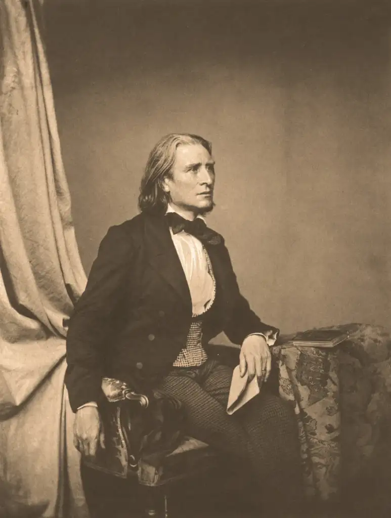 A sepia-toned portrait of a man with long hair seated beside a table. He wears a formal suit with a bow tie and holds papers in one hand. The background features a draped curtain and a patterned cloth on the table.