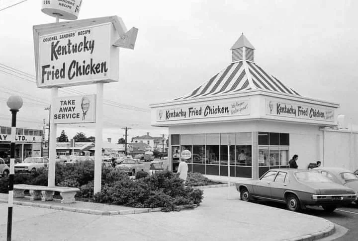 Black and white photo of a vintage Kentucky Fried Chicken with a striped roof. A prominent sign reads "Colonel Sanders' Recipe." Several cars and people are visible in the parking lot. The atmosphere is retro, suggesting a mid-20th century setting.