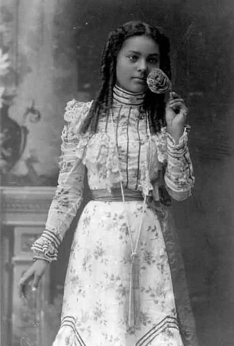 A vintage black and white photograph of a woman in a floral dress holding a seashell near her face. She has long, curly hair and is standing in a room with ornate decor.