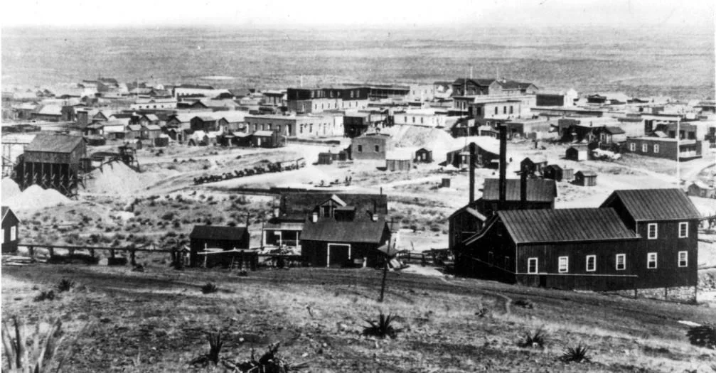 Black and white image of a historical mining town with wooden buildings scattered across a barren landscape. Dirt roads are visible, and a rugged terrain surrounds the town. A few tall structures and sparse vegetation can be seen.