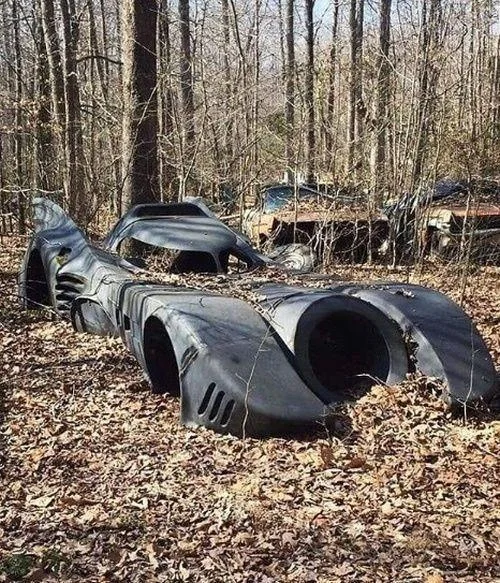 A rusted, abandoned Batmobile shell sits among dry leaves and trees in a forest setting, surrounded by other old, weathered cars.