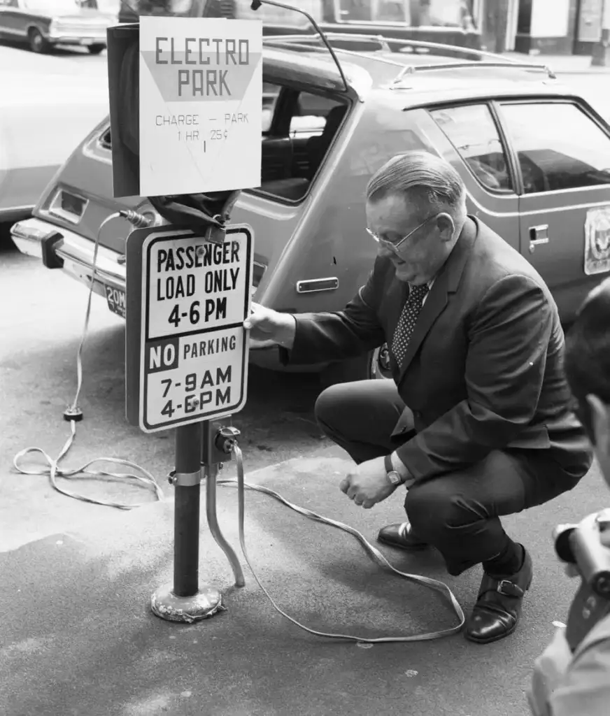 A man kneels next to a car on a city street, examining an "Electro Park" setup. Signs indicate parking rules and charging fees. A cable connects the car to the station.