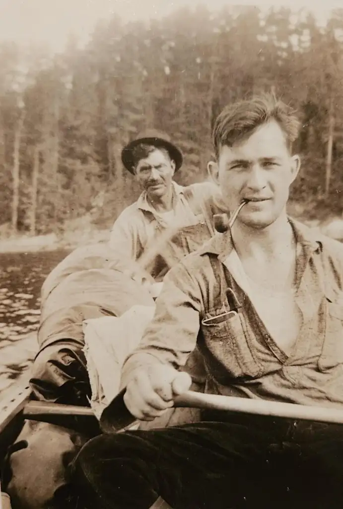 Two men are in a small boat on a lake, paddling. The man in the foreground is holding an oar and has a pipe in his mouth. Both are wearing casual shirts. Blurred trees are visible in the background. The image is in sepia tone.