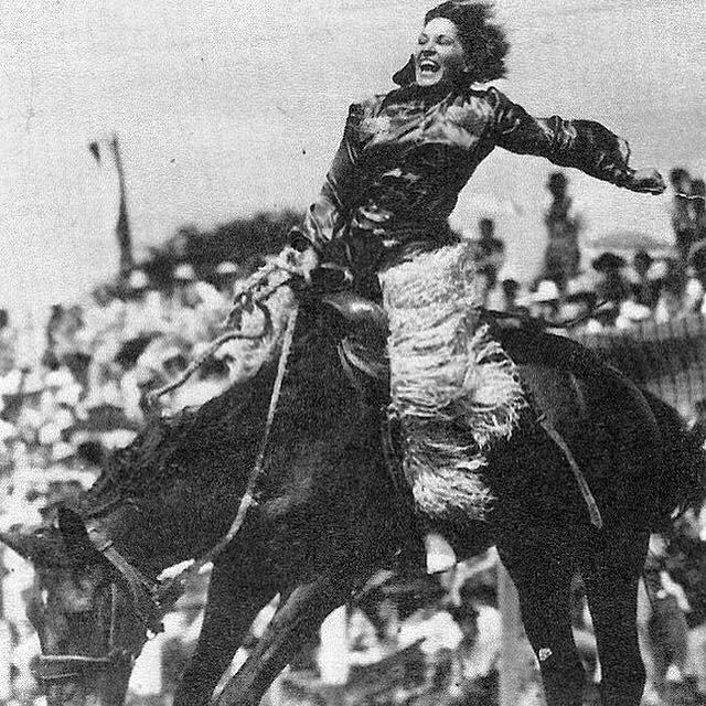 A woman rides energetically on a moving horse during a rodeo, wearing a shiny shirt and fringed chaps. A crowd of spectators is visibly watching in the background. The image is in black and white, capturing a dynamic moment.