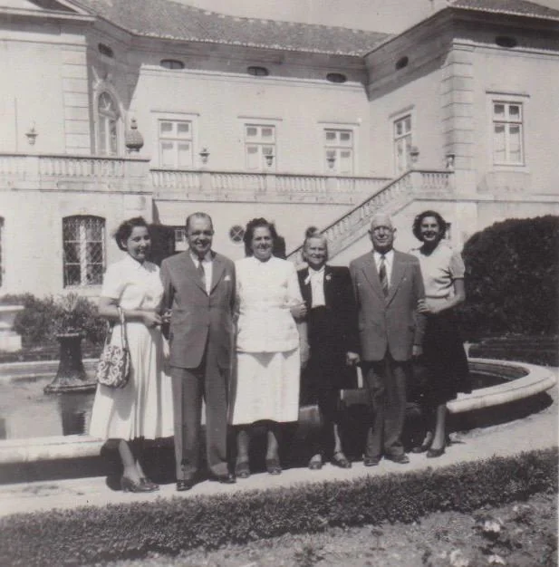 A group of six people, four men and two women, stand arm in arm in front of a large, elegant building with a fountain. The setting appears formal, with everyone dressed in suits and dresses, likely from a past era.