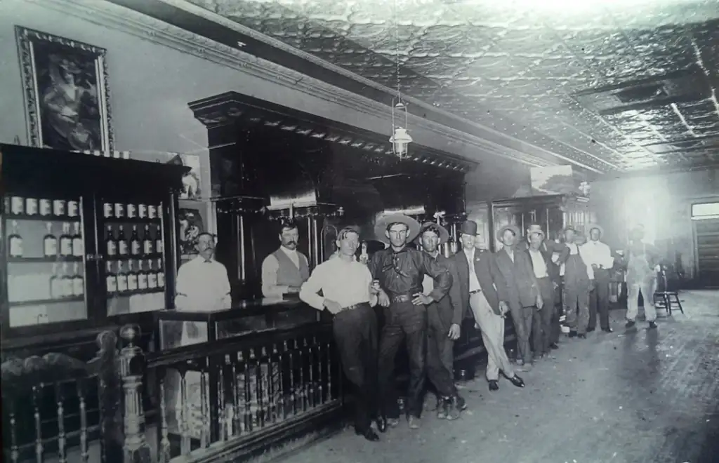 Black and white photo of a vintage bar scene with nine men posing. They stand along a long wooden bar, dressed in early 20th-century attire. The bar is stocked with bottles, and an ornate ceiling and framed picture are visible above.