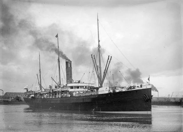 A vintage black and white photograph of a steamship sailing on water. Smoke billows from its tall funnel, and multiple masts rise above the deck. The ship's hull is dark, and there are several people visible on board.