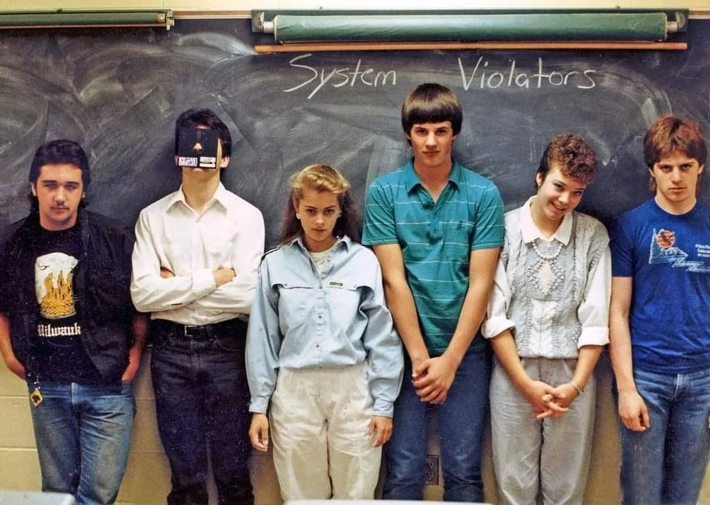 A group of six people stand in front of a chalkboard with "System Violators" written on it. They appear to be in a classroom, wearing casual 1980s attire, with expressions ranging from serious to playful.