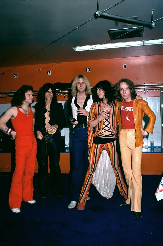 Five people in colorful 1970s attire stand in a locker room-like setting. They wear vibrant outfits, including striped pants, bell-bottoms, and a Ferrari T-shirt. A blue carpet and numbered lockers are visible in the background.