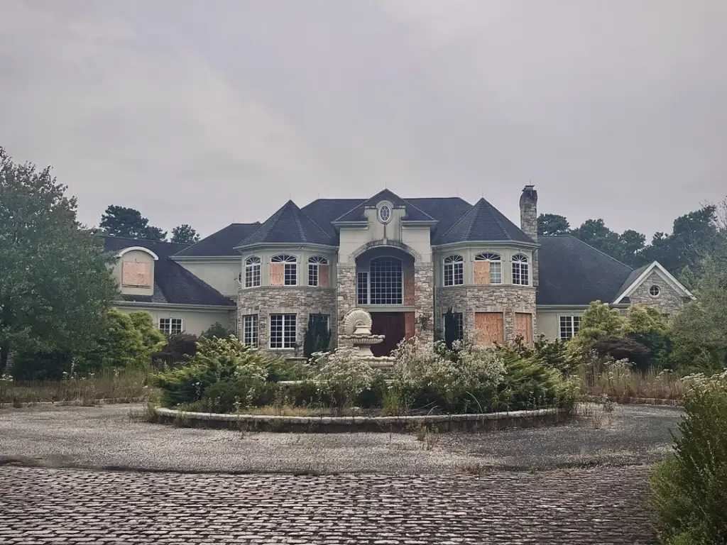 Abandoned mansion with boarded-up windows, surrounded by overgrown vegetation. The two-story building features a circular driveway and fountain at the center, with a cloudy sky in the background.