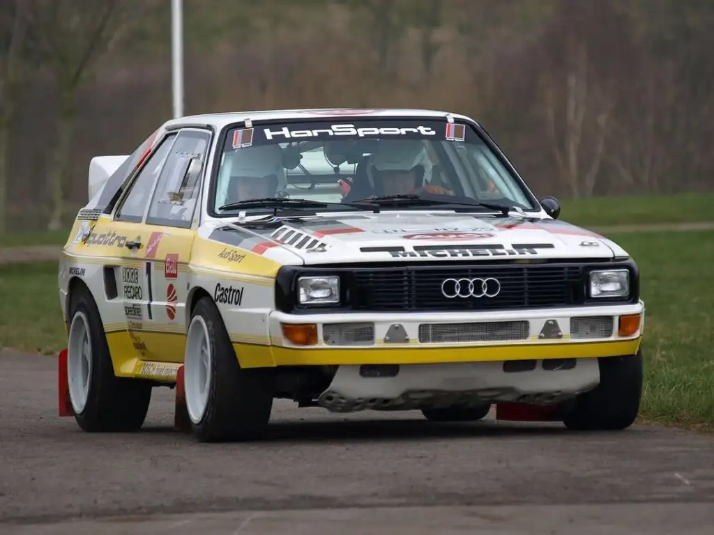 A classic Audi rally car with a yellow and white livery and various sponsor logos navigates a curve on a paved road. The car has a distinctive boxy shape and large headlights, with trees and grass visible in the background.