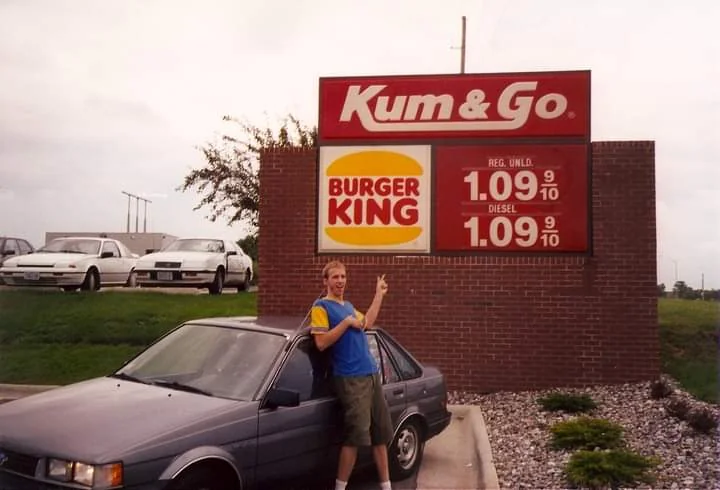 A person stands next to a parked car, pointing at a sign for "Kum & Go" and "Burger King" with fuel prices listed as $1.09 per gallon for both regular unleaded and diesel. Several cars are visible in the background.