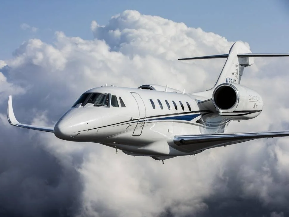 A sleek, white private jet flies through the sky, surrounded by fluffy white clouds. The aircraft is angled slightly upwards, suggesting ascent, with a blue stripe along its side. The sky is mostly cloudy with patches of sunlight.