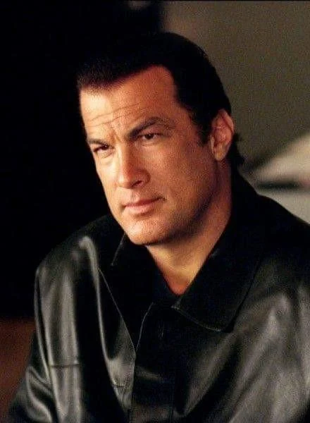 A person with short dark hair, wearing a black leather jacket, sits indoors. They have a serious expression, and the background is softly blurred.