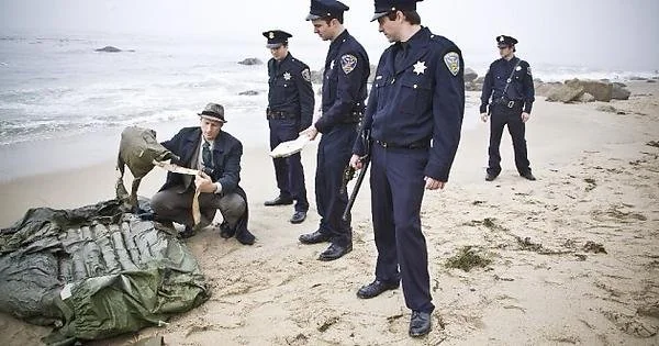 Five people, including a person in a suit and four in police uniforms, stand on a foggy beach examining a large, green, canvas object spread on the sand. The ocean and rocks are visible in the background.