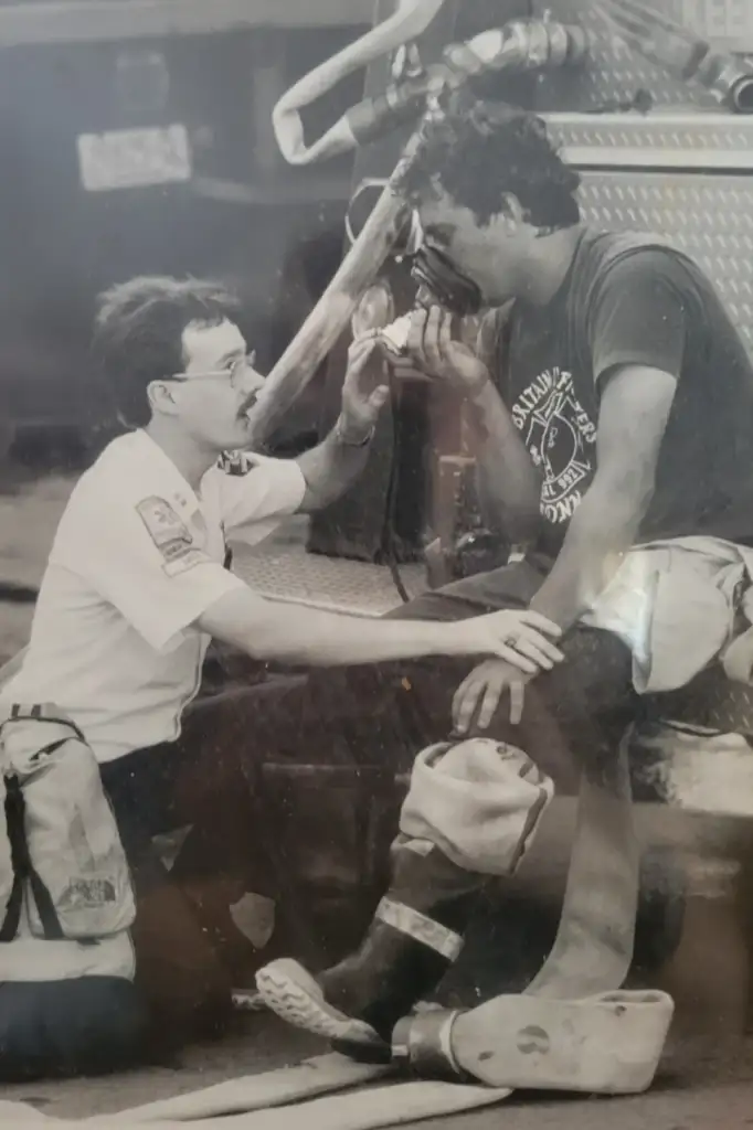 A firefighter sits on a firetruck bumper, wearing gear and holding an oxygen mask. Another person, in an EMT uniform, kneels beside him, offering support. They are engaged in a focused and concerned interaction.