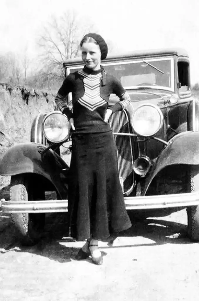 A woman in a long skirt and patterned blouse stands confidently in front of an old-fashioned car, with her hands on her hips. The background shows a rural landscape with bare trees under a clear sky.