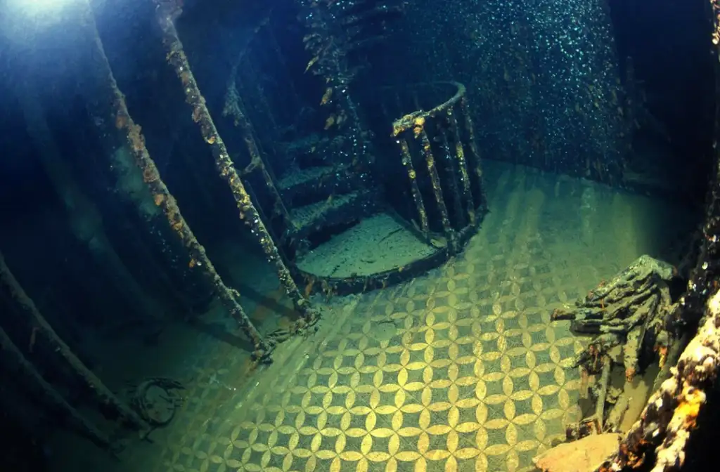 Underwater image of a sunken ship’s interior showing a circular spiral staircase and detailed tiled floor. Rust and marine life cover metal structures, creating a mysterious and tranquil atmosphere beneath the sea.