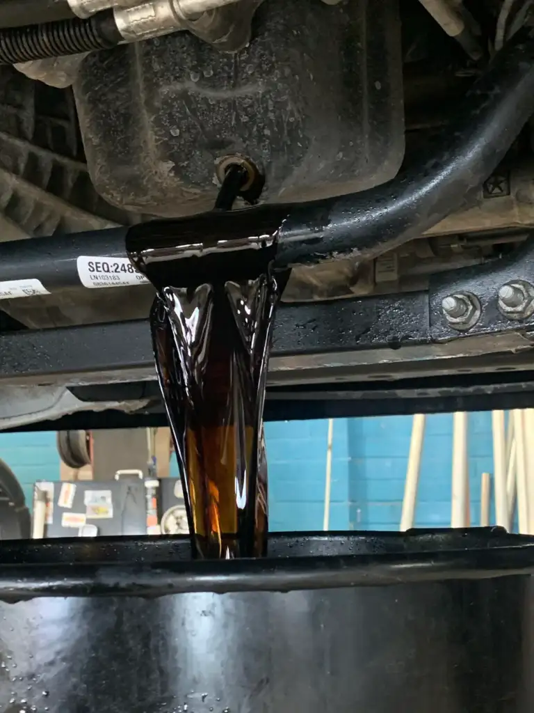 Close-up of thick, dark oil pouring from a vehicle's underside into a black container below. The background features a blurred interior with pipes and a blue wall.