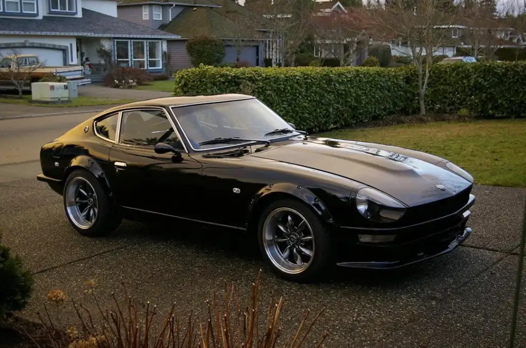 A shiny black classic sports car is parked on a suburban street with neatly trimmed hedges and houses in the background. The car features sleek curves and polished metallic wheels, reflecting the soft natural light.