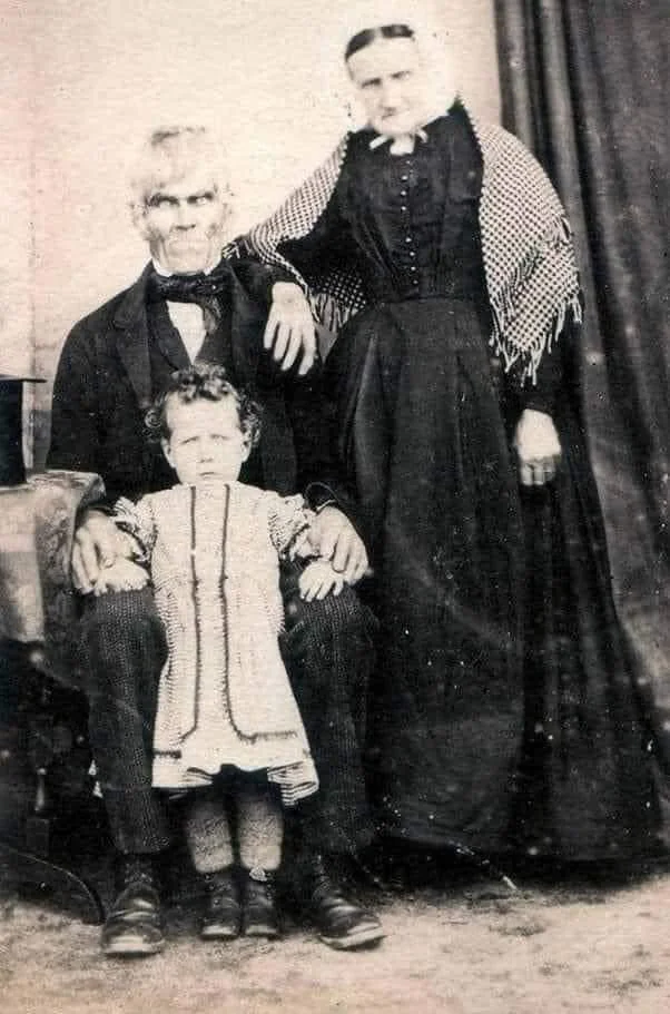 A vintage black and white photo of an older couple with a child. The man is seated, wearing a suit, with the child sitting on his lap. The woman stands beside them, dressed in a long gown and shawl. The backdrop is a simple curtain.