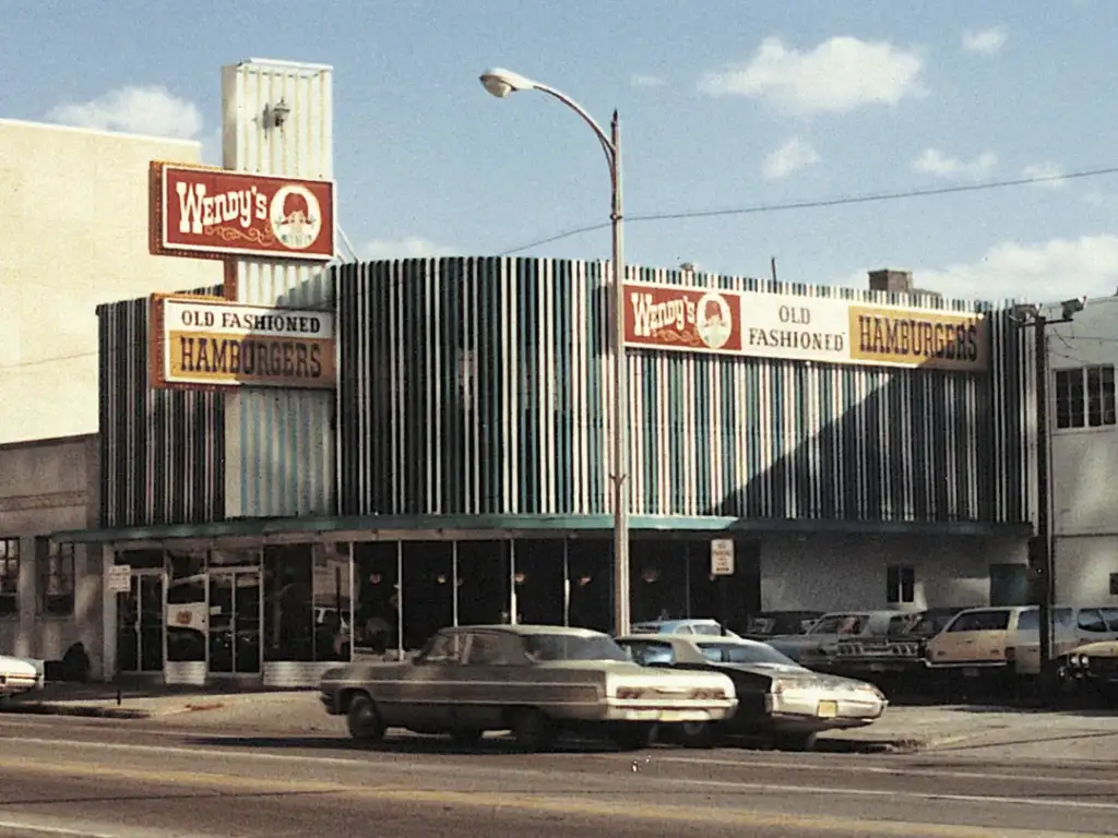 Vintage photo of a Wendy's restaurant with "Old Fashioned Hamburgers" signs. The building has a distinctive striped design, with a sign on the roof. Classic cars are parked in front. The sky is partly cloudy.