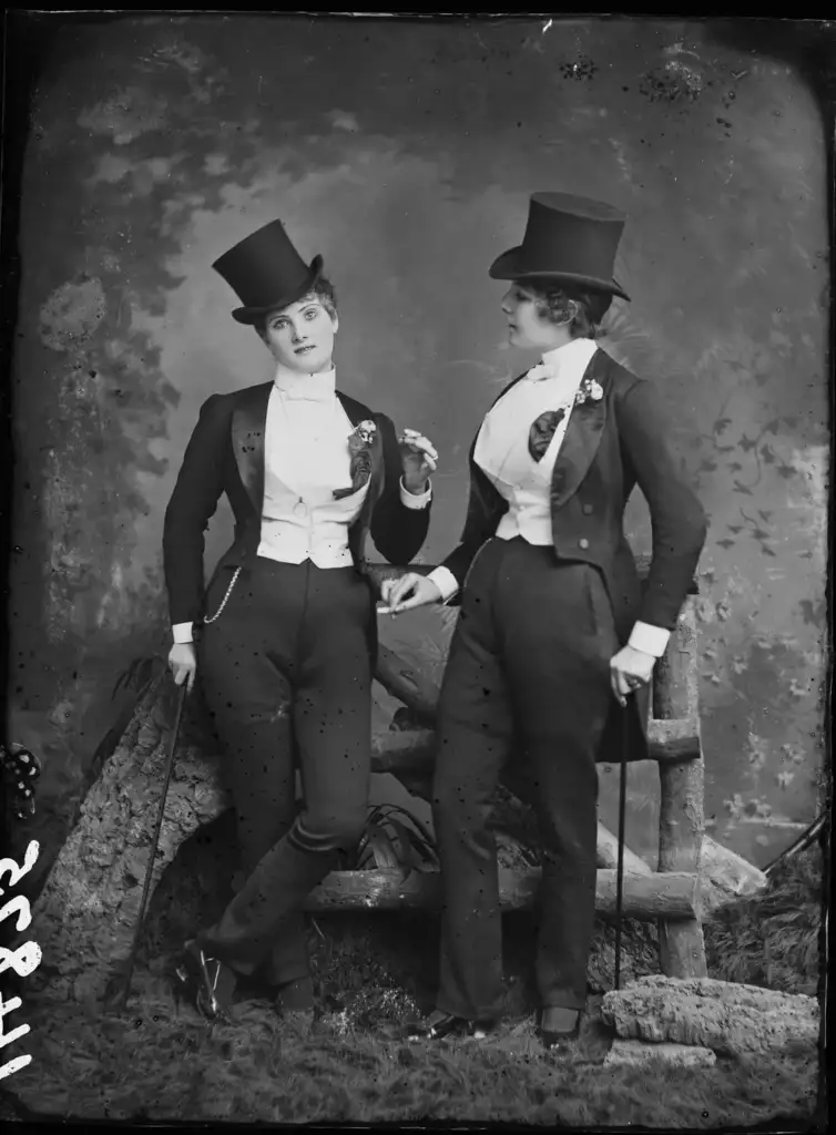 Two women in vintage attire, wearing top hats, tailcoats, and holding canes, pose with confident expressions. One leans against a wooden fence, while the other stands nearby. The backdrop features a textured, natural setting.