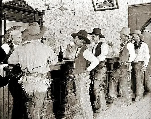 A sepia-toned image of cowboys in a saloon. Several men in hats and vests stand at a wooden bar, drinking and chatting. A bartender is serving them. The scene is set in the Old West, with vintage decor and attire.