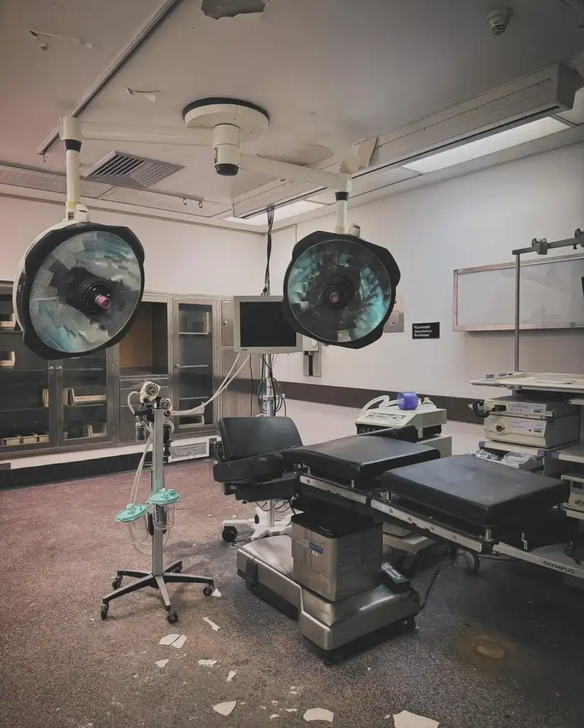 An empty hospital operating room with overhead surgical lights, an operating table, and various medical equipment. The room has a monitor, cabinets, and a stand with face masks. The floor has some debris and the ceiling shows wear.