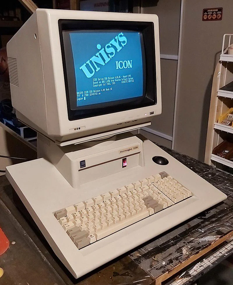 A vintage Unisys Icon computer with a CRT monitor and beige keyboard sits on a wooden table. The screen displays the Unisys logo and some text. The setup is in a dimly lit room with shelves and various items in the background.